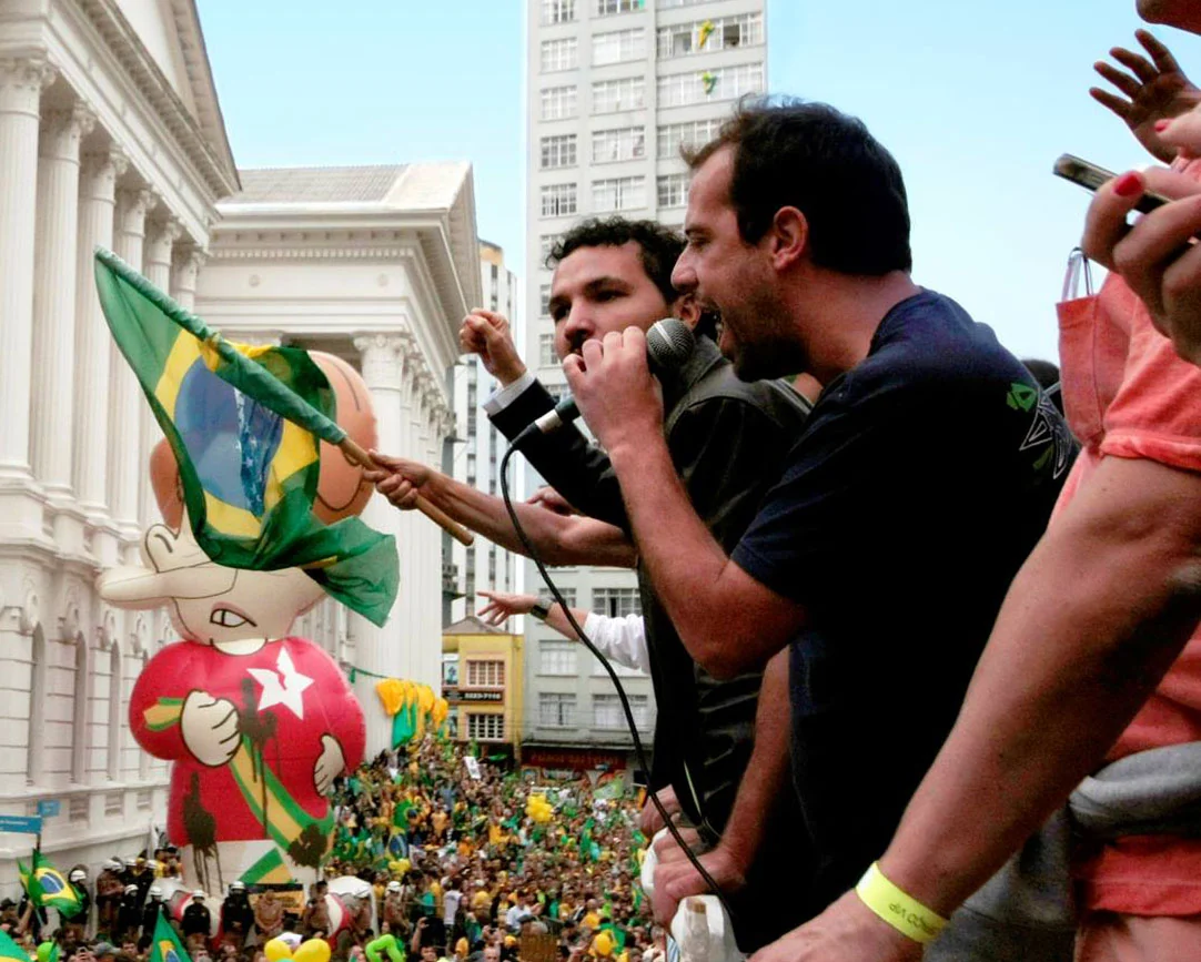 Eder Borges ao microfone em manifestação no centro de Curitiba, diante de multidão com bandeiras do Brasil